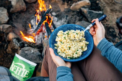 Seated by a campfire, a person holds a blue bowl of macaroni and cheese with herbs-one of many tasty camping recipes-while rocks and a bag of Kettle brand chips rest nearby.