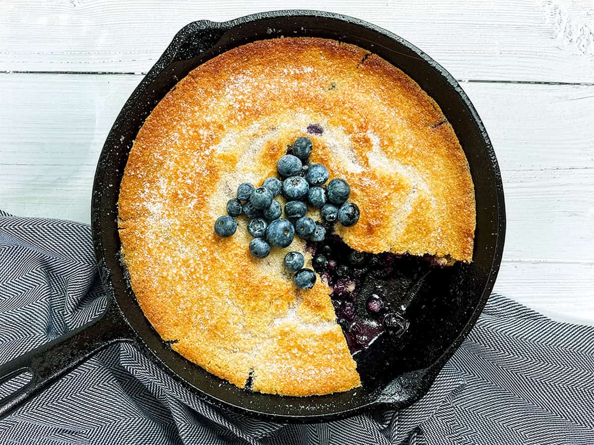 A skillet blueberry cobbler sits in a cast iron pan, topped with fresh blueberries. One slice is missing, revealing the juicy blueberry filling inside.
