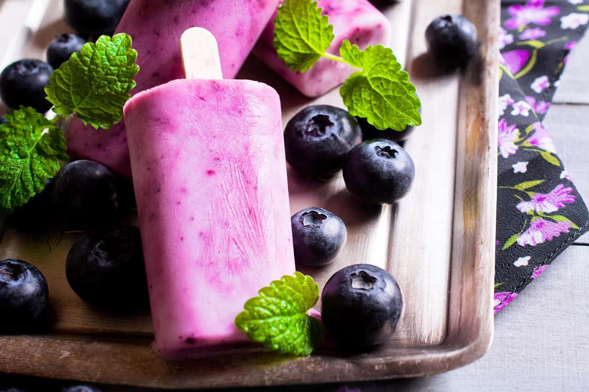 Pink berry and blueberry popsicles on a metal tray, surrounded by fresh blueberries and mint leaves, with a floral napkin at the side.
