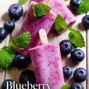 Three blueberry popsicles rest on a wooden surface, surrounded by fresh blueberries and mint leaves.