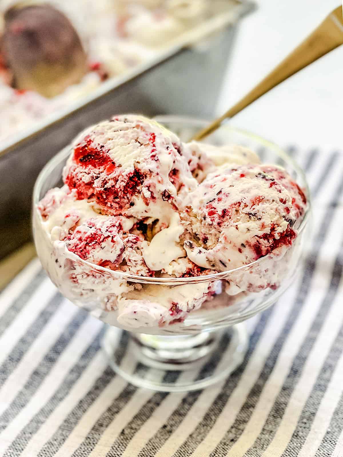 A glass bowl filled with scoops of Berry No Churn Ice Cream, placed on a striped cloth with a metal pan of ice cream in the background.
