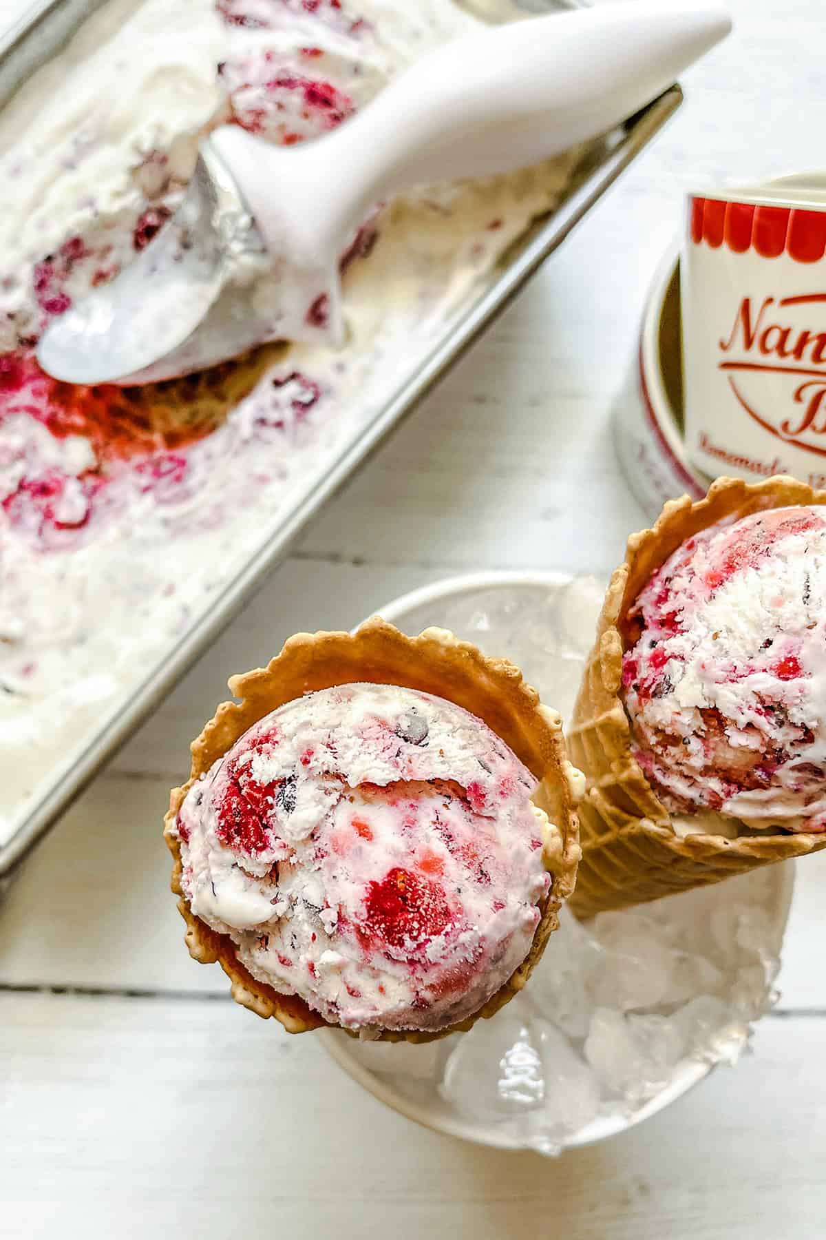 Two waffle cones with scoops of Berry No Churn Ice Cream rest on ice beside a tub of ice cream and an ice cream scooper on a white surface.
