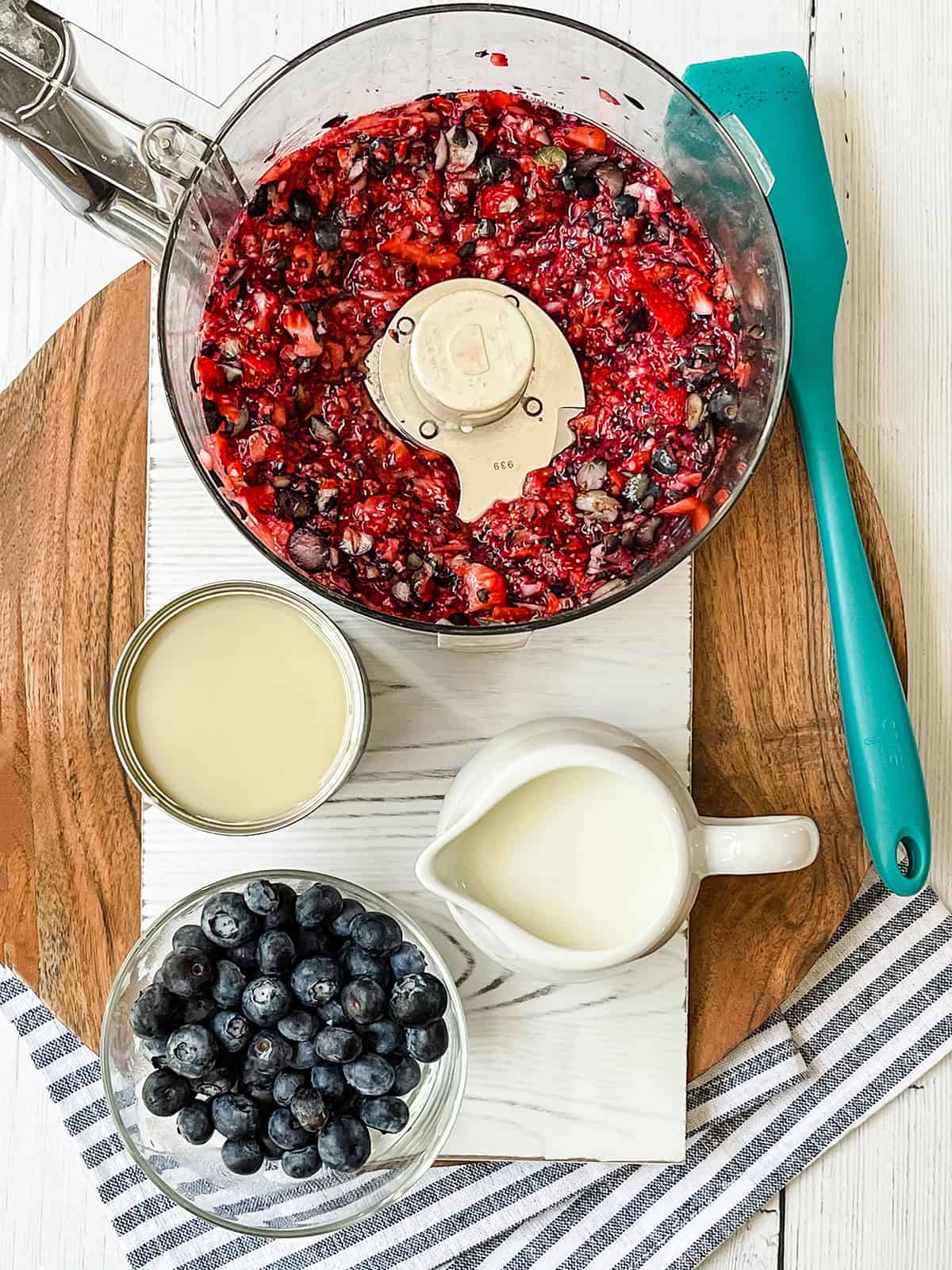 A food processor with chopped berries, a bowl of blueberries, a pitcher of cream, and a can of condensed milk are arranged on a round wooden board-perfect ingredients for Berry No Churn Ice Cream-with a blue spatula and striped cloth.
