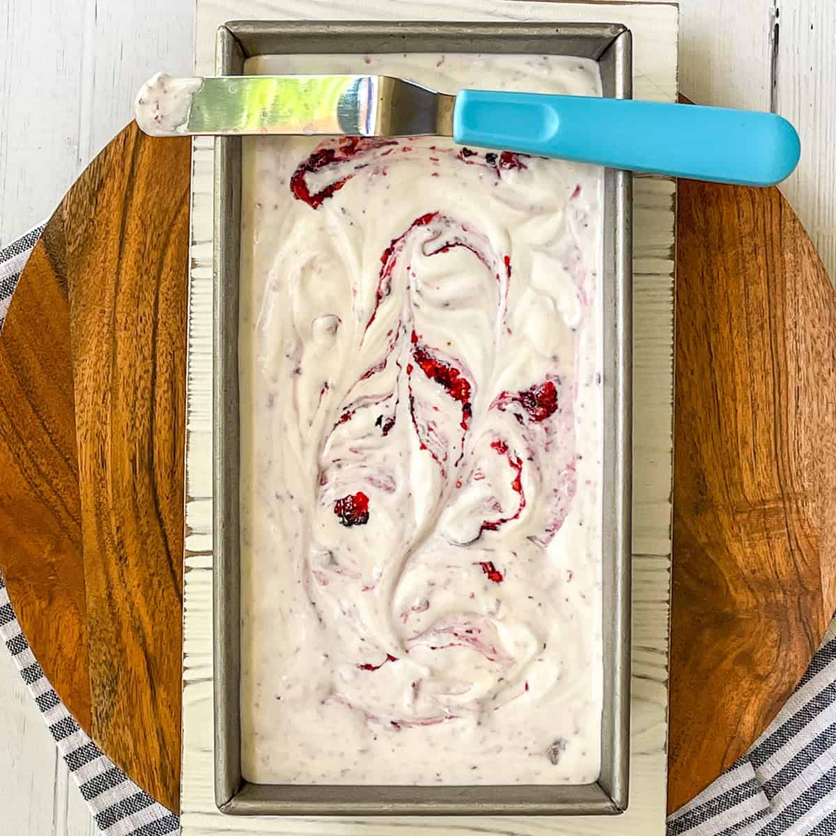 A rectangular metal pan filled with creamy Berry No Churn Ice Cream swirled with red fruit, topped with a blue-handled spatula, sits on a wooden board.