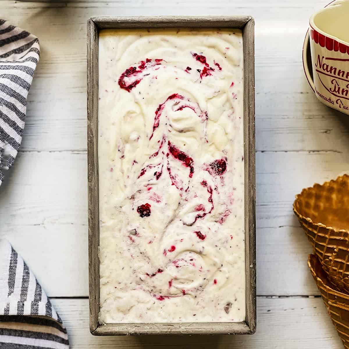 A rectangular metal pan filled with Berry No Churn Ice Cream-vanilla ice cream swirled with raspberry sauce-sits next to a waffle cone, a striped towel, and a mug on a white wooden surface.