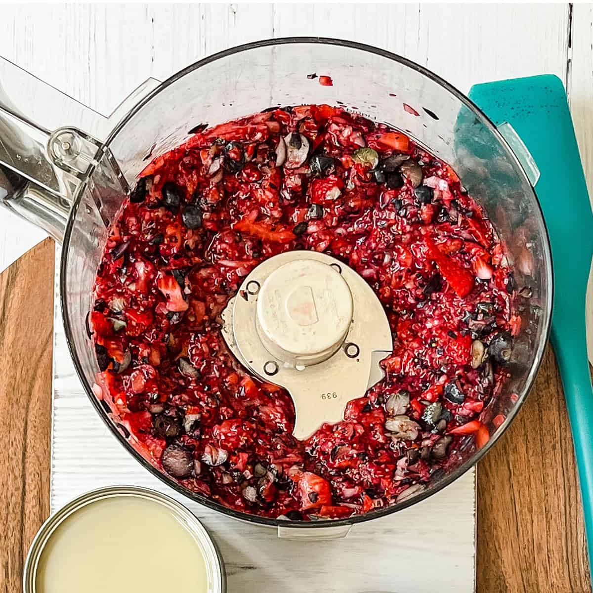 A food processor bowl containing a chopped mixture of red berries and dark beans sits on a wooden cutting board, ready to be transformed into Berry No Churn Ice Cream, with a blue spatula and an open can nearby.