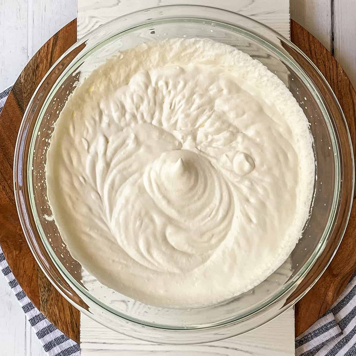 A glass bowl filled with freshly whipped cream sits on a wooden board atop a white surface, ready to be folded into homemade Berry No Churn Ice Cream.