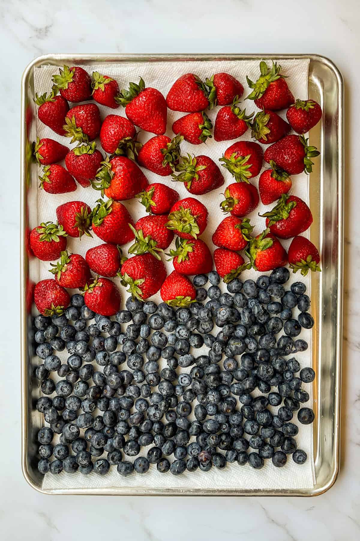 A baking tray lined with paper towels holds fresh strawberries on the top half and blueberries on the bottom half, arranged in neat rows-perfect ingredients for a vibrant berry trifle.