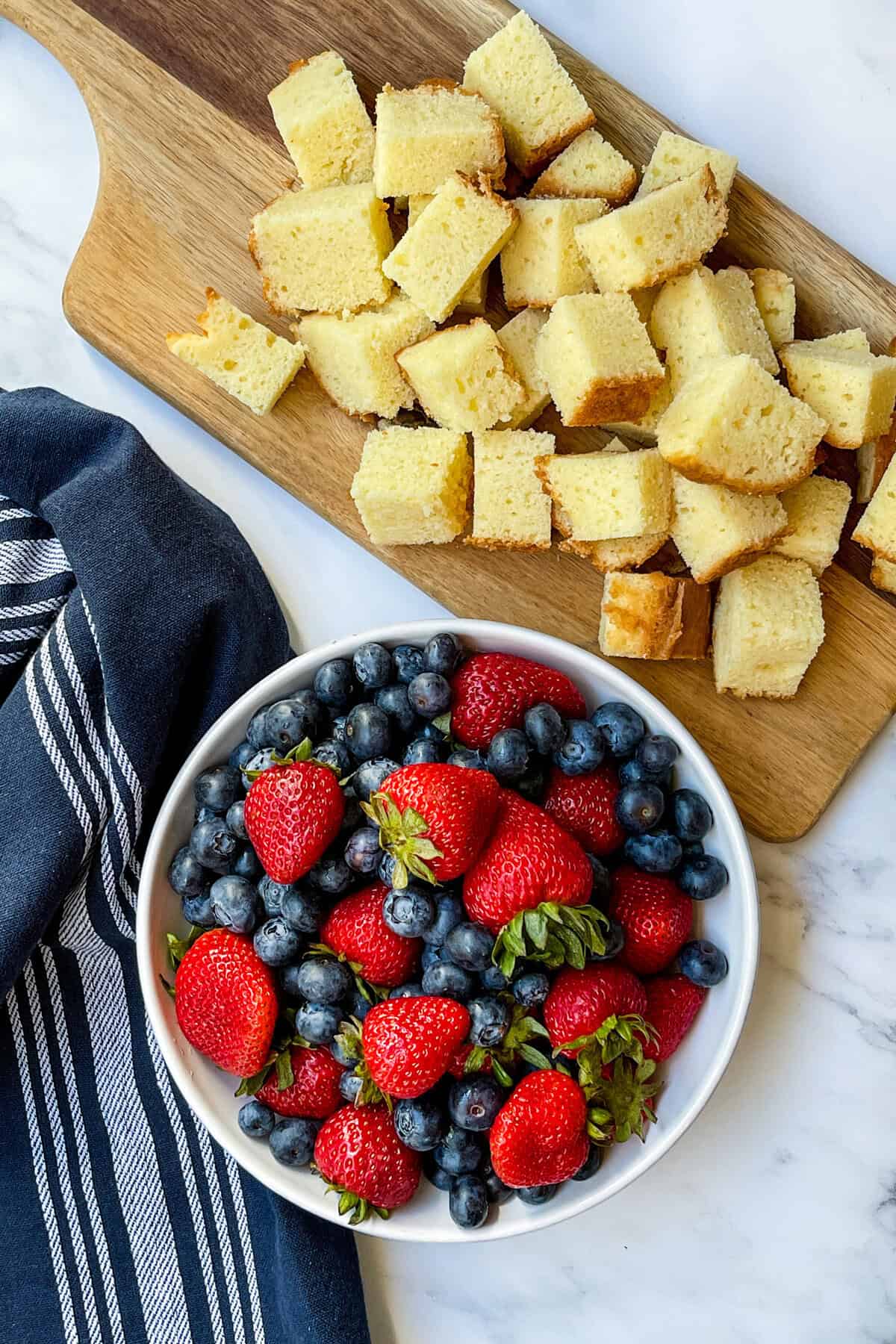 A white bowl filled with fresh strawberries and blueberries sits next to cubed pieces of yellow cake, ready for assembling a berry trifle, all arranged on a wooden cutting board beside a navy striped cloth on a marble surface.