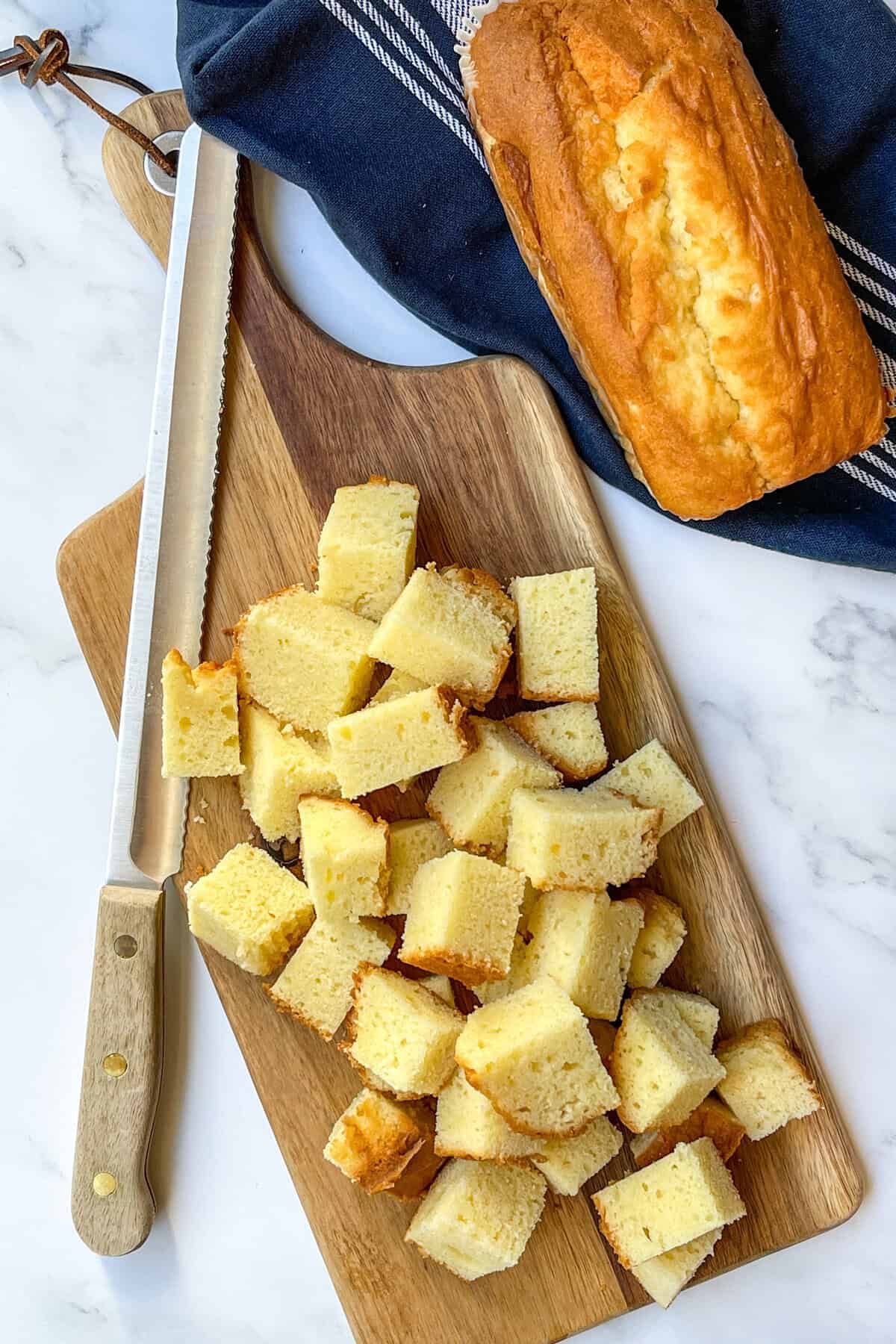 A loaf of pound cake on a wooden cutting board, partially sliced into cubes for a berry trifle, with a serrated knife beside it. A navy cloth is in the background on a white marble surface.