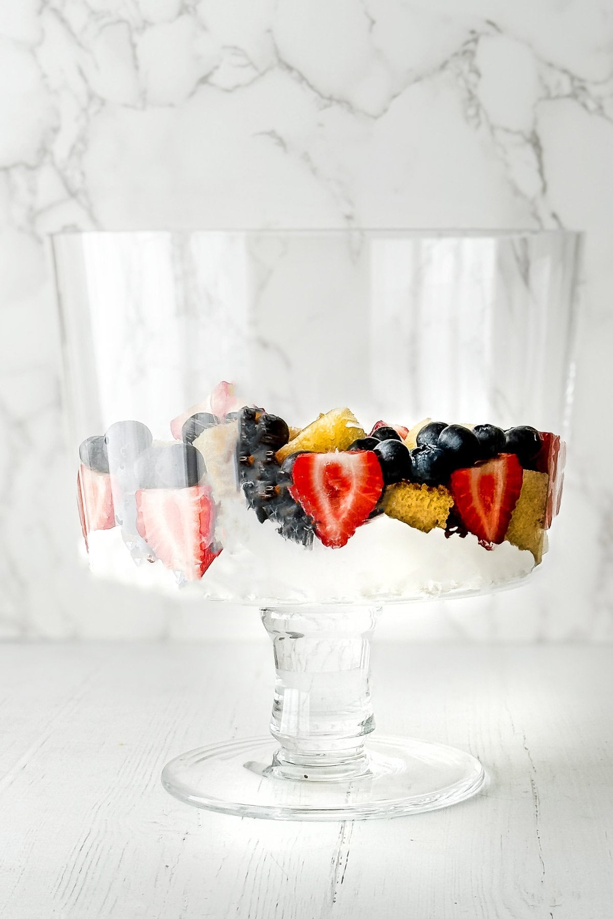 A clear glass trifle dish filled with sliced strawberries, blueberries, blackberries, and pieces of yellow cake creates a stunning berry trifle, arranged in a ring pattern on a white surface with a marble background.