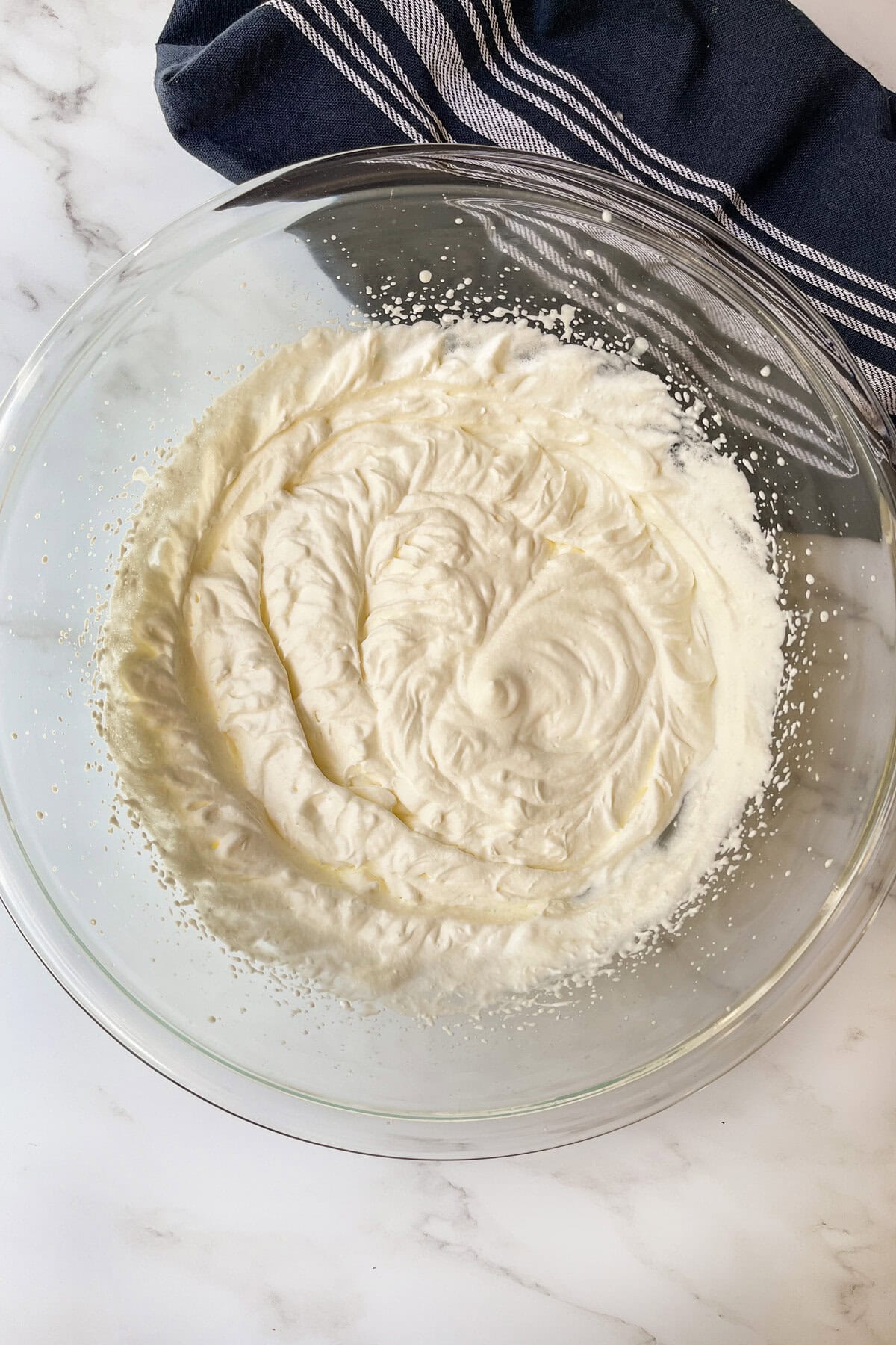 A glass bowl filled with freshly whipped Chantilly cream sits on a white marble surface, ready to top a berry trifle, with a dark blue striped kitchen towel partially visible in the background.