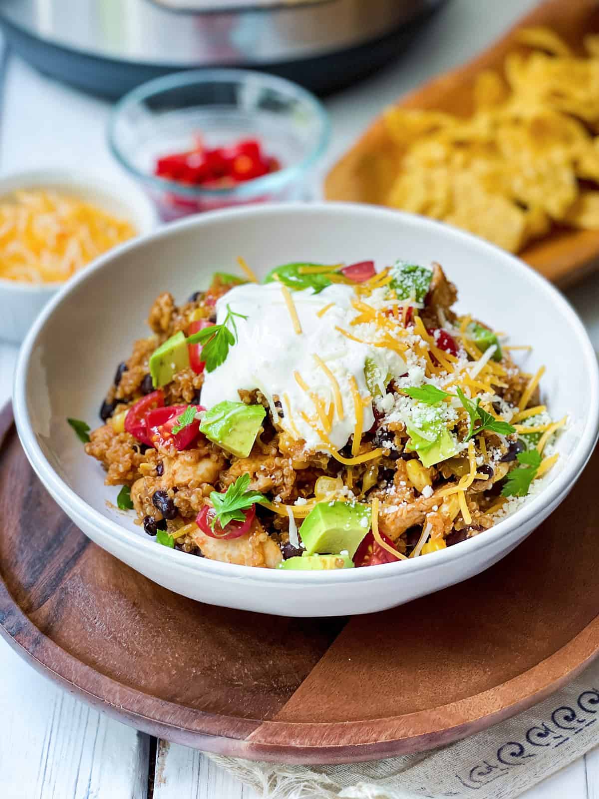 A bowl of chicken enchilada quinoa bowls topped with sour cream, shredded cheese, avocado, and tomatoes, with chips, extra cheese, and sliced peppers in the background.