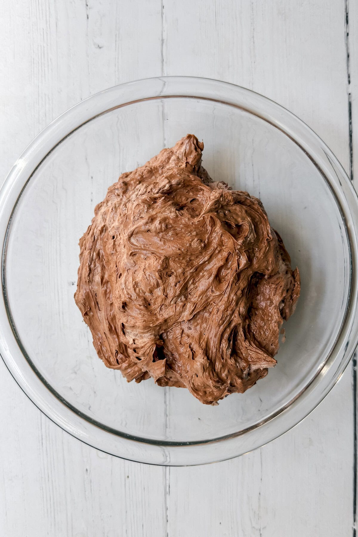 A large ball of chocolate bread dough, perfect for making a decadent chocolate pie, sits in a clear glass bowl on a white wooden surface.