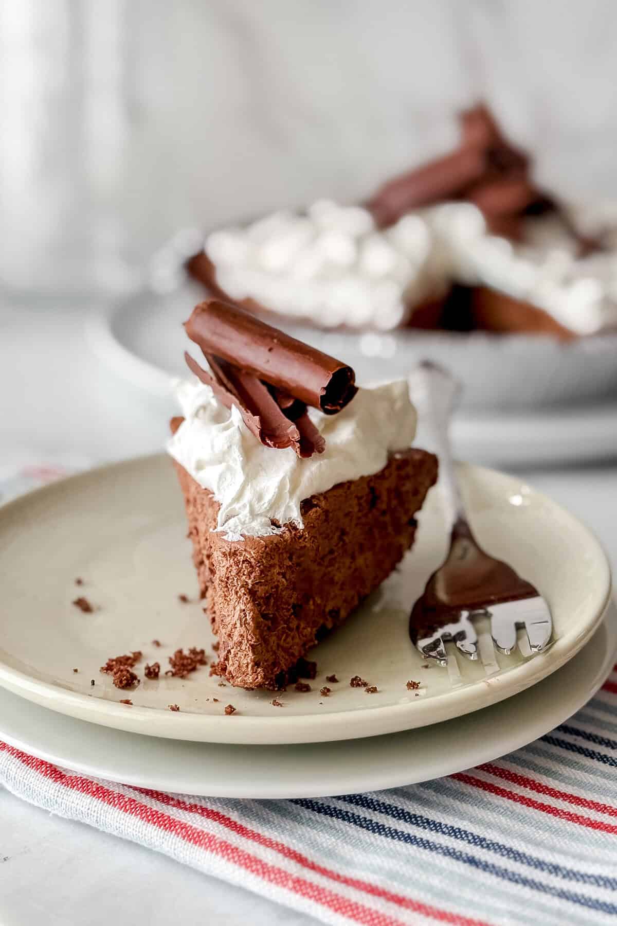 A slice of chocolate pie mousse topped with whipped cream and chocolate curls sits on a plate with a fork and crumbs, while the rest of the chocolate pie is blurred in the background.