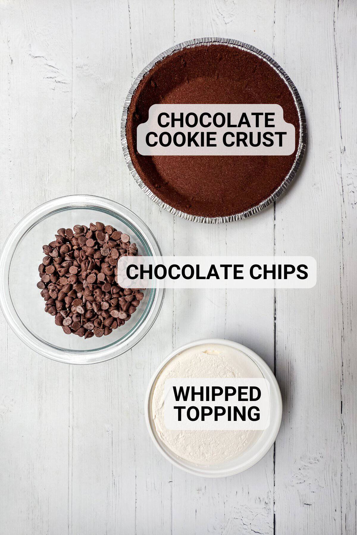Top-down view of three labeled dessert ingredients for chocolate pie: a chocolate cookie crust in a foil pan, a glass bowl of chocolate chips, and a container of whipped topping, all on a white wooden surface.