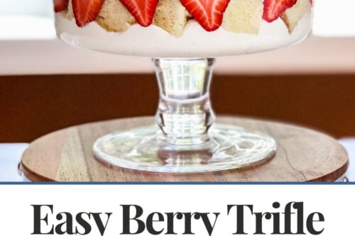 A berry trifle in a glass dish features layers of whipped cream, strawberries, blueberries, and cake cubes, displayed on a table. Below, a cutting board holds extra cake cubes beside a bowl of fresh berries.