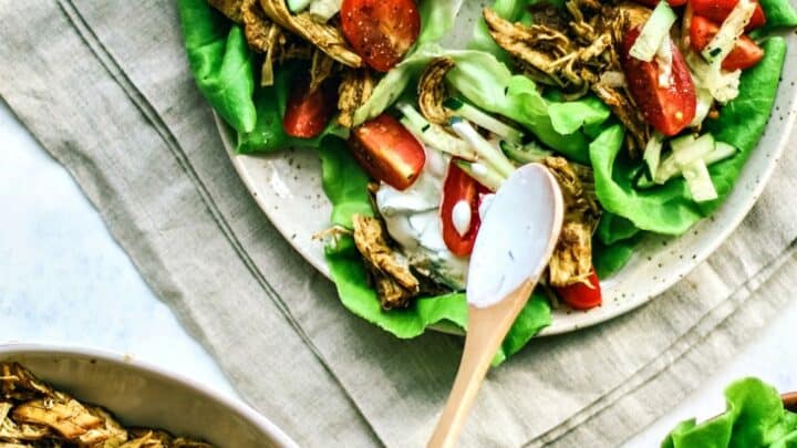 Three lettuce wraps filled with shredded chicken, tomatoes, cucumber, and sauce on a plate; a hand adds sauce with a spoon. Perfect for Summer 20 minute dinners, with fresh ingredients visible in bowls nearby.