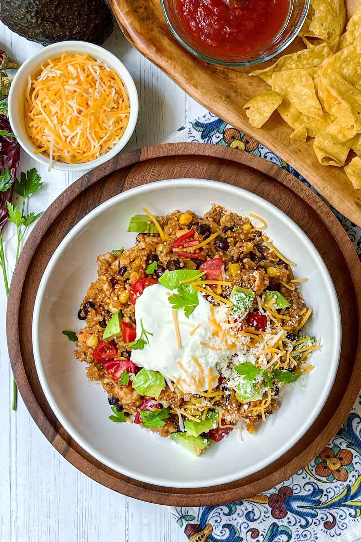 A bowl of Mexican quinoa topped with sour cream, shredded cheese, avocado, tomatoes, and herbs sits beside tortilla chips and salsa on a colorful tablecloth—perfect alongside instant pot chicken enchilada casserole.