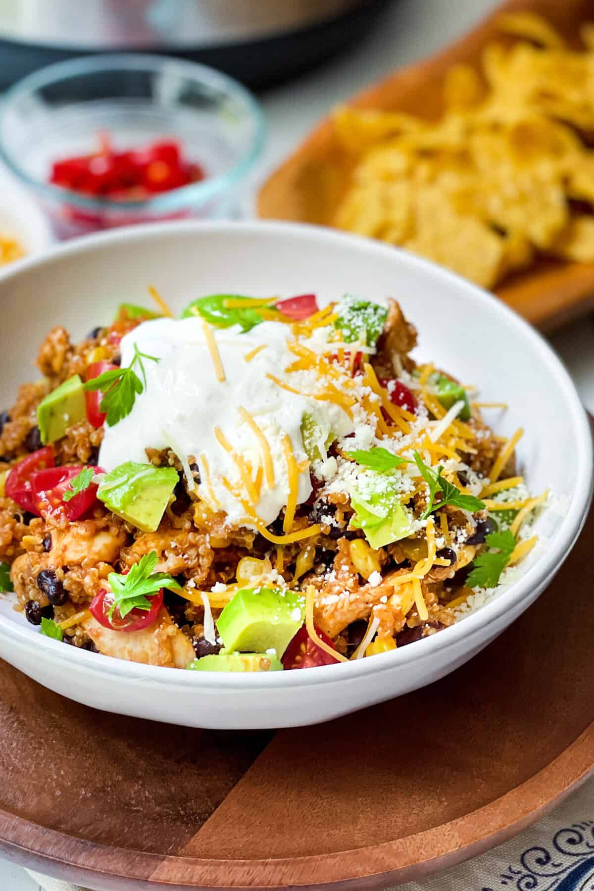 A bowl of Mexican-inspired rice topped with sour cream, shredded cheese, diced avocado, tomatoes, parsley, and crumbled cheese, reminiscent of an instant pot chicken enchilada casserole, with corn chips and extra toppings in the background.