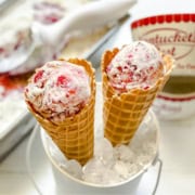 Two waffle cones filled with Berry No Churn Ice Cream are nestled in a white bucket of ice, with a tub of ice cream and a scoop in the background.