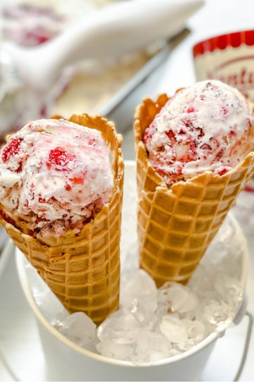 Two waffle cones filled with Berry No Churn Ice Cream are nestled in a white bucket of ice, with a tub of ice cream and a scoop in the background.
