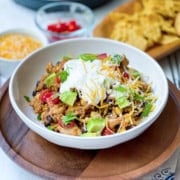 A bowl of rice, black beans, avocado, tomato, shredded cheese, and sour cream sits on a wooden tray alongside chips-perfect sides for a hearty chicken enchilada quinoa bowls; extra toppings are visible in the background.
