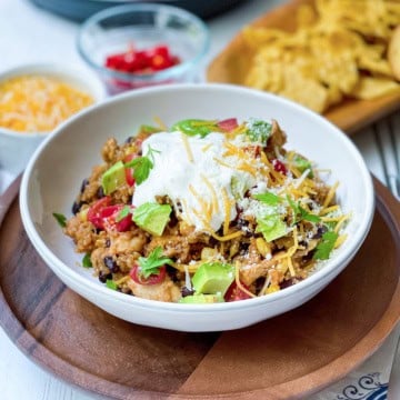 A bowl of rice, black beans, avocado, tomato, shredded cheese, and sour cream sits on a wooden tray alongside chips-perfect sides for a hearty chicken enchilada quinoa bowls; extra toppings are visible in the background.