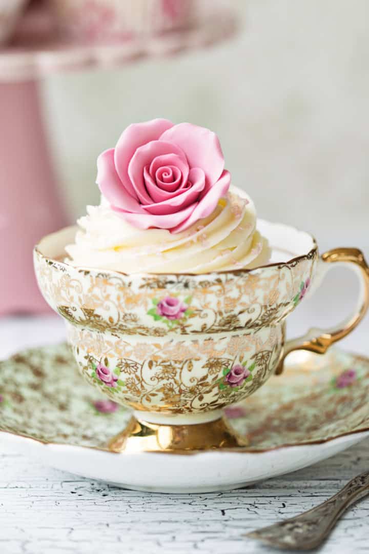 A decorative cupcake topped with a pink rose sits in an ornate floral teacup on a matching saucer, ready to delight as one of your afternoon tea sweets, with a fork beside it on a white textured surface.