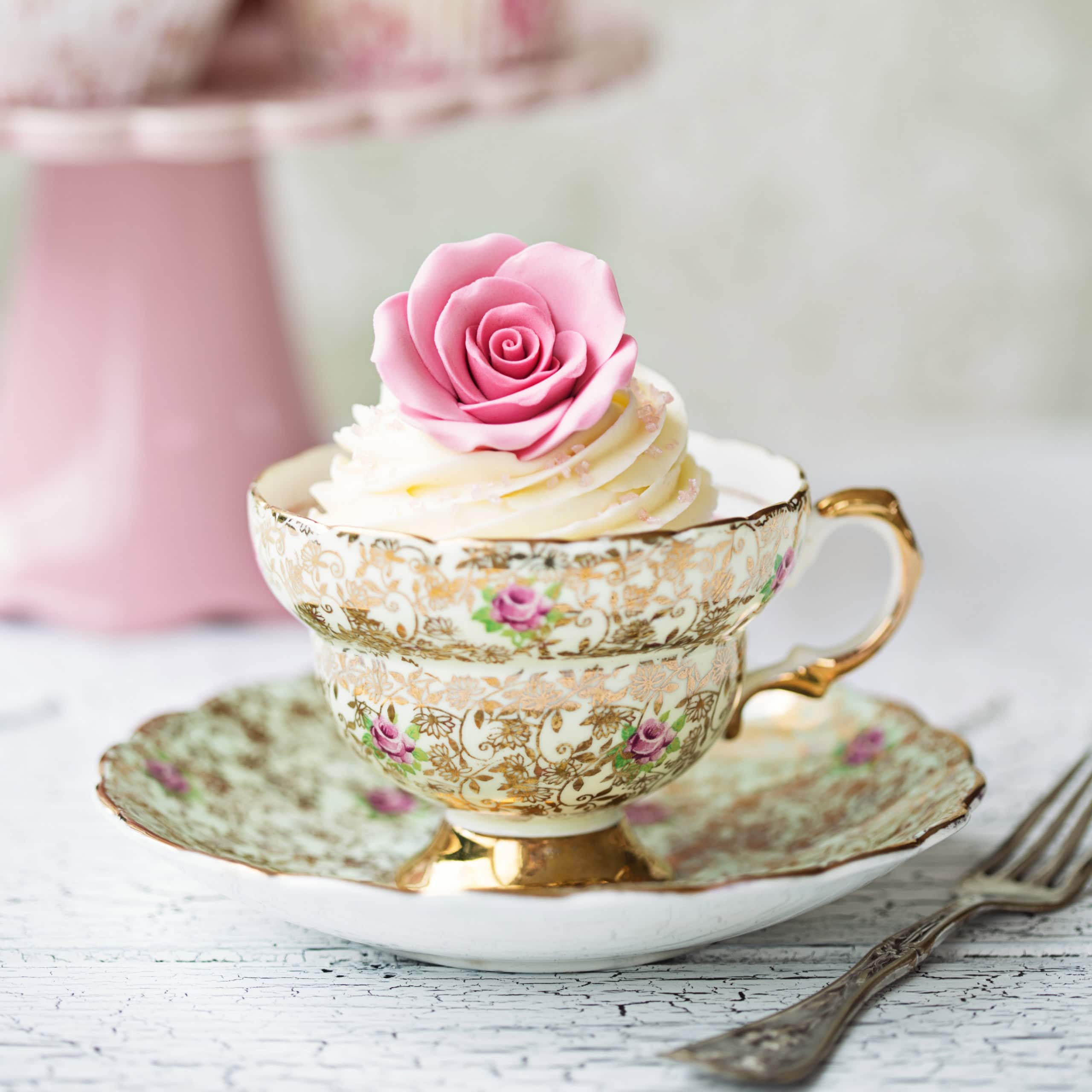 A decorative cupcake topped with a pink rose sits in an ornate floral teacup on a matching saucer, ready to delight as one of your afternoon tea sweets, with a fork beside it on a white textured surface.