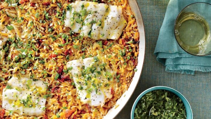 A skillet with baked fish fillets over orzo pasta, garnished with herbs, sits beside three glasses of white wine, a bowl of herbs, and a halved lemon on a blue tablecloth—perfect for July One Pot Dinners.