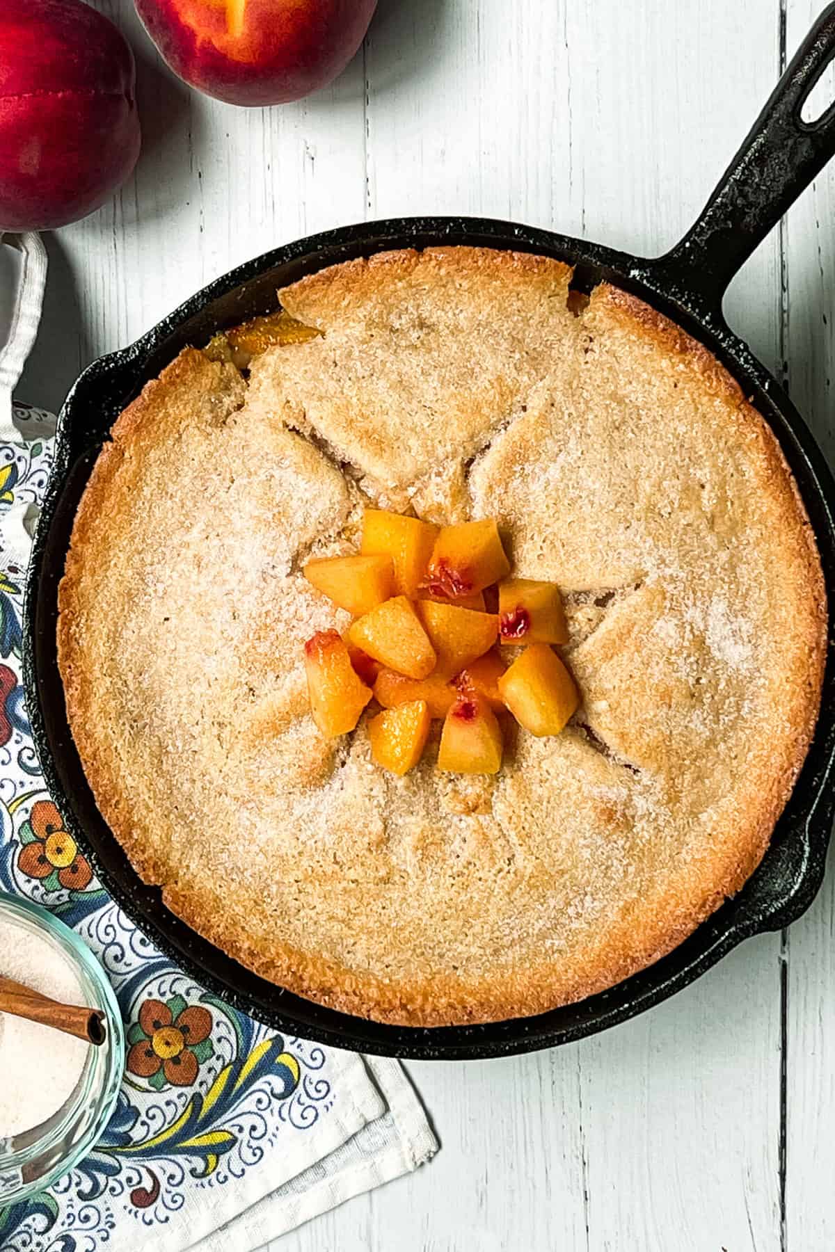 Freshly baked peach cobbler, dusted with sugar, and topped with chopped fresh peaches in a cast iron skillet.