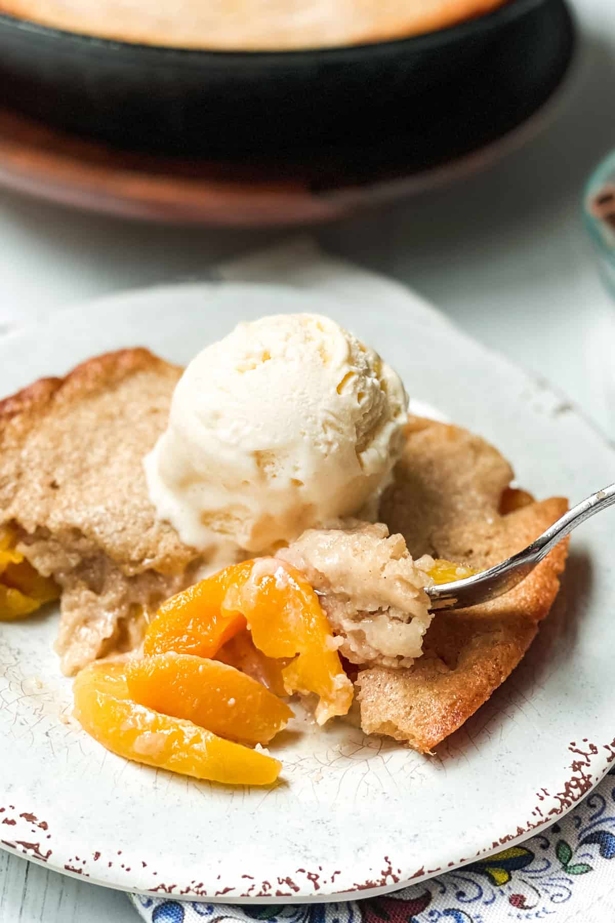 Serving of skillet peach cobbler with melting vanilla ice cream on a plate with a fork.