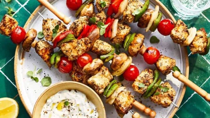 Grilled chicken and vegetable skewers with tomatoes, mushrooms, peppers, and onions on a plate, served with tzatziki sauce and lemon wedges—perfect for Summer Mediterranean Diet Dinners on a green tiled surface.