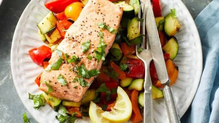A plate with a cooked salmon fillet on top of chopped roasted vegetables, garnished with herbs and served with lemon wedges, a fork, and a knife—a perfect choice for summer Mediterranean Diet dinners.