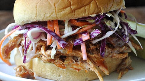 A pulled pork sandwich with coleslaw on a white plate, garnished with a pickle spear, makes for a tasty addition to your no-cook dinners, served on a rustic wooden surface.