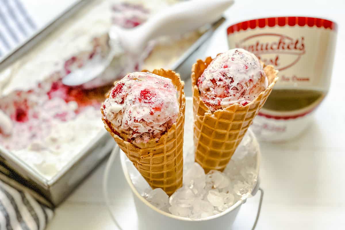Two waffle cones filled with Berry No Churn Ice Cream are placed in a bucket of ice, with a tub of ice cream and scoop in the background.