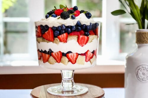 A berry trifle dessert in a glass bowl, featuring whipped cream, blueberries, strawberries, and sponge cake, topped with fresh berries and mint leaves, displayed on a wooden stand near a window with soft natural light.