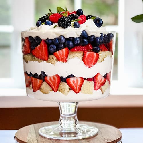A berry trifle dessert in a glass bowl, featuring whipped cream, blueberries, strawberries, and sponge cake, topped with fresh berries and mint leaves, displayed on a wooden stand near a window with soft natural light.