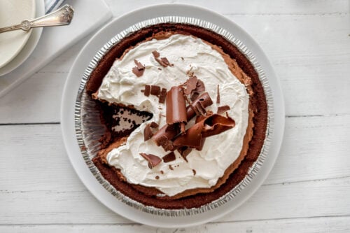 A decadent chocolate pie with a graham cracker crust, topped with whipped cream and chocolate curls, sits on a white plate. One slice has been removed, showcasing the rich chocolate filling inside.