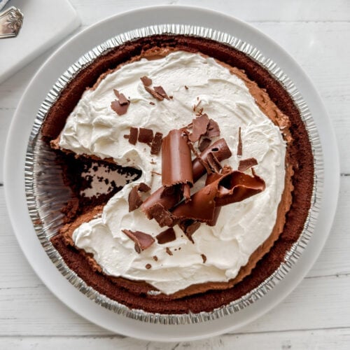 A decadent chocolate pie with a graham cracker crust, topped with whipped cream and chocolate curls, sits on a white plate. One slice has been removed, showcasing the rich chocolate filling inside.