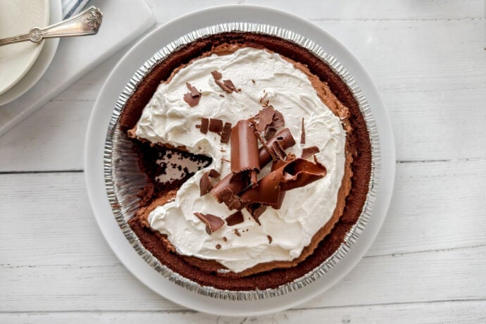 A decadent chocolate pie with a graham cracker crust, topped with whipped cream and chocolate curls, sits on a white plate. One slice has been removed, showcasing the rich chocolate filling inside.