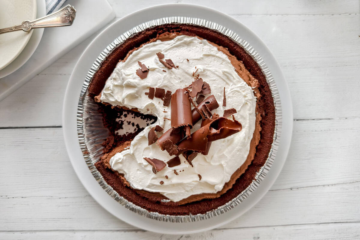 A decadent chocolate pie with a graham cracker crust, topped with whipped cream and chocolate curls, sits on a white plate. One slice has been removed, showcasing the rich chocolate filling inside.