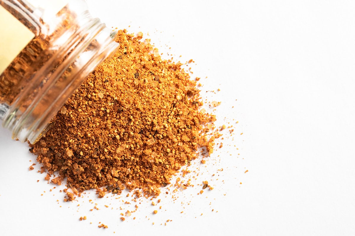 A glass jar tipped on its side spills homemade dry rub spice mixture onto a white surface.