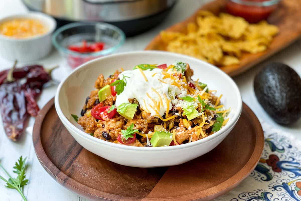 A bowl of instant pot chicken enchilada casserole topped with sour cream, shredded cheese, avocado, and herbs sits on a wooden plate. In the background are chips, grated cheese, sliced peppers, and an avocado.