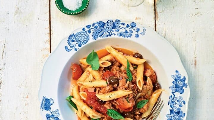 A plate of penne pasta with tomato sauce, olives, and basil sits on a white wooden table&mdash;perfect for Summer Mediterranean Diet Dinners&mdash;accompanied by a fork, a glass of white wine, and small bowls of salt and pepper.