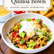 A bowl of chicken enchilada quinoa, inspired by classic chicken enchilada casserole, topped with avocado, sour cream, shredded cheese, and veggies sits on a wooden board with the recipe title above.
