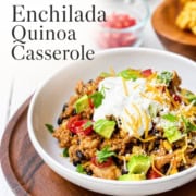 A bowl of chicken enchilada casserole quinoa topped with sour cream, shredded cheese, avocado, and herbs, with a side of chopped tomatoes in the background.