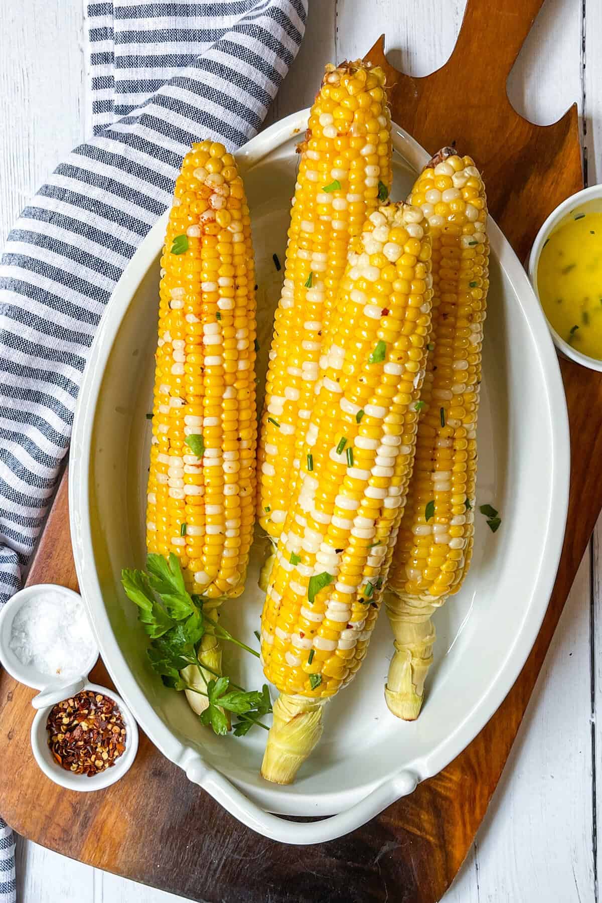 Three ears of grilled corn on the cob garnished with herbs rest on an oval white dish, accompanied by small bowls of salt, red pepper flakes, and a yellow sauce.