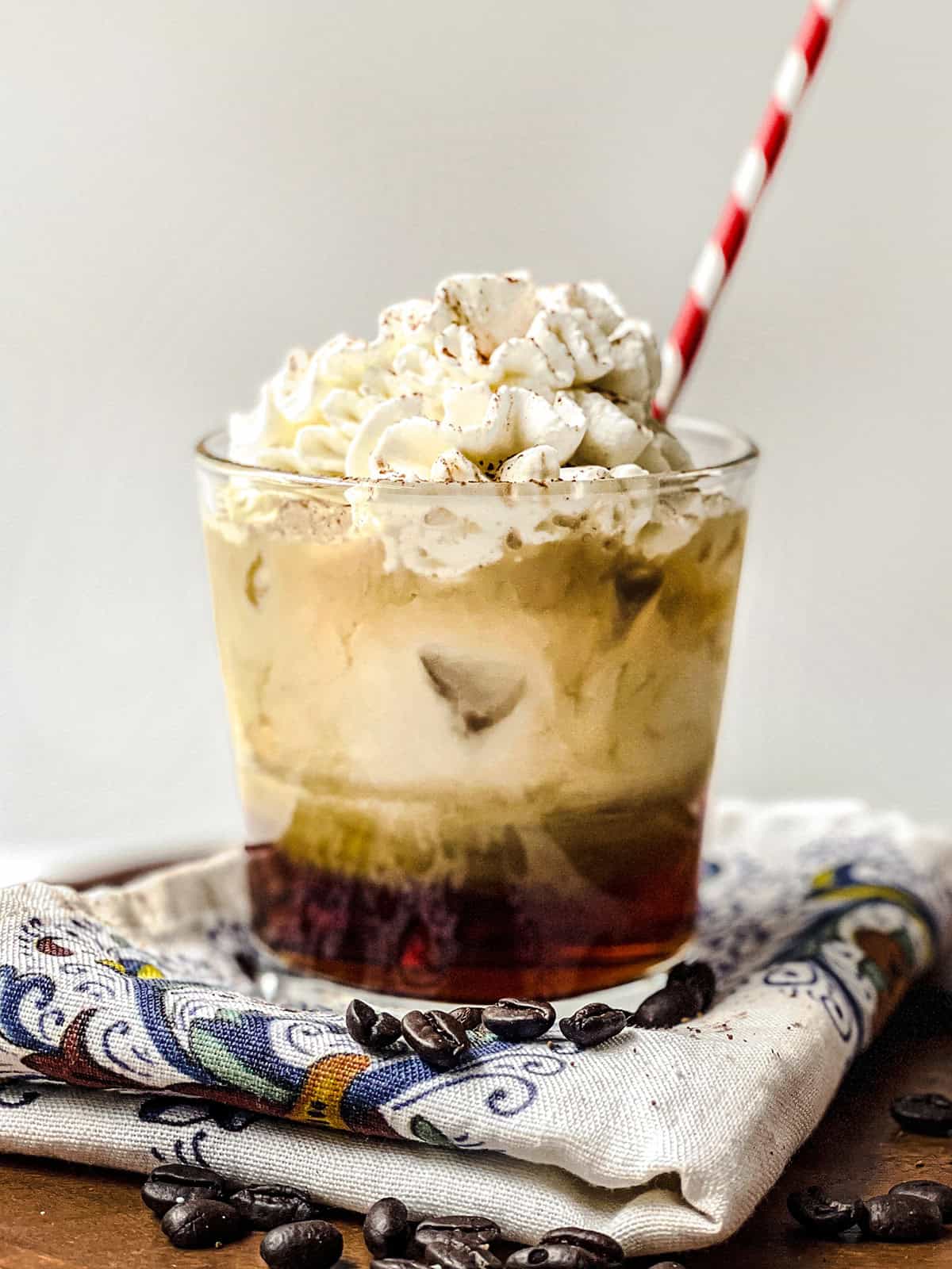 A glass of iced vanilla latte topped with whipped cream and a striped straw, placed on a cloth napkin with coffee beans scattered around.